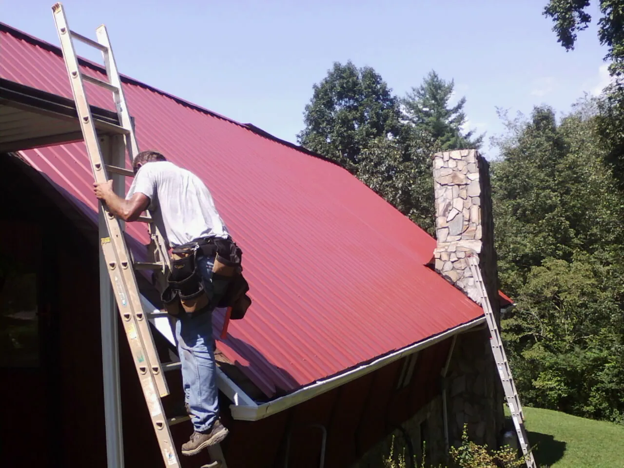 Roofer on a ladder beside a red metal roof and stone chimney — Soffit & Fascia Repair by Roofing Brothers in Mesquite